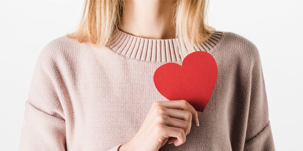 Person hält ein rotes Papierherz vor die Brust. Darstellung kann auf Herzgesundheit und Blutdruckmessung hinweisen, Hintergrund ist weiß und schlicht.